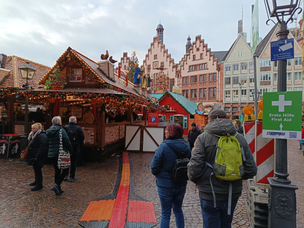 Eine Gruppe von Menschen spaziert auf einem Kopfsteinpflasterweg neben einem Weihnachtsmarkt in Nürnberg, Deutschland, mit Laternenmasten, Texttafeln und Gebäuden mit Fenstern im Hintergrund unter einem bewölktem Himmel.