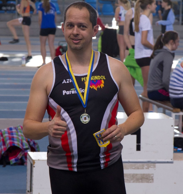 Ein Mann in schwarzem Jersey und Medaille steht, während einige Menschen im Hintergrund trainieren.