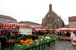 Ein belebter Markt in Nürnberg, Deutschland, mit farbenfrohen Obst- und Gemüseständen, Menschen mit Taschen und aufgestellten Zelten, umgeben von Gebäuden und einem Uhrenturm im Hintergrund unter einem sichtbaren Himmel.