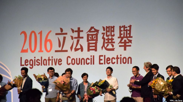 Eine Gruppe von Menschen auf einer Bühne mit Blumensträußen, mit einem Publikum darunter und einem Hintergrundbild mit Text und Logo für die Wahl zum Legislativrat 2016.