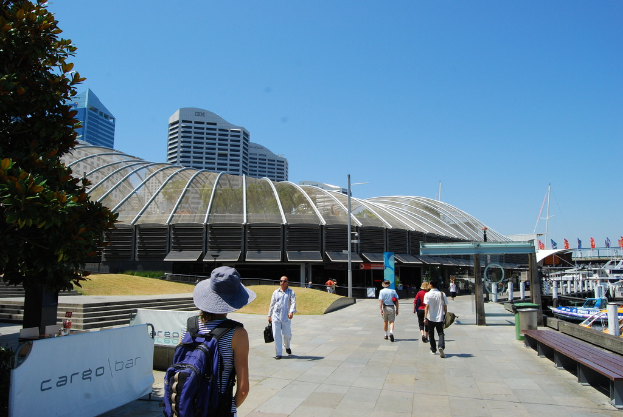 Menschen auf einem Gehweg mit Mülltonnen, Bänken, Holzlatten und Metallstangen auf der rechten Seite, Pfosten, Bäume und Metalllattenzaun mit Bannern auf der linken Seite und Gebäude im Hintergrund.