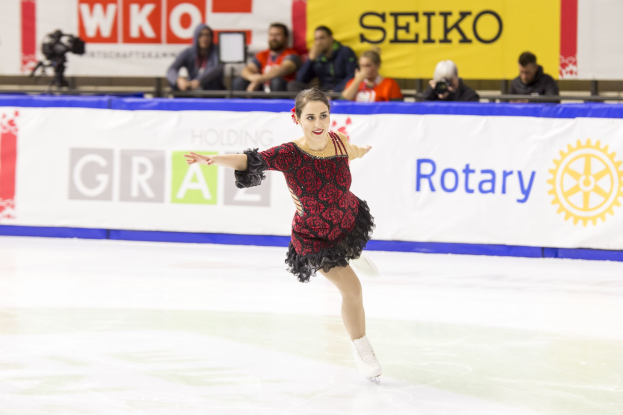 Eine Frau in einem roten und schwarzen Kleid, die auf einer Eisbahn während der Weltmeisterschaften im Eiskunstlaufen skatet, mit Zuschauern und einem Fotografen im Hintergrund.