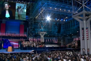 Eine große Menschenmenge sitzt einer Bühne mit einem Rednerpult, einem Mikrofon, Flaggen, einem Bildschirm und Lichtausrüstung zugewandt, was auf eine Präsidentschaftsdebatte hinweist.