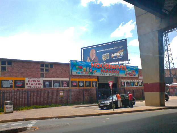 Eine Straße mit einer Säule und einem Auto auf der rechten Seite, ein Pfahl mit einer weißen und roten Tafel, ein Auto mit zwei Männern daneben, ein Müllcontainer auf der linken Seite, braune Gebäude mit Plakaten im Hintergrund, eine blaue Tafel mit Text oben auf einem Gebäude, eine blaue Werbetafel, ein Stromtransformator und ein rotes Objekt auf der rechten Seite und ein bewölkter Himmel darüber.