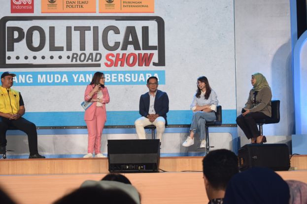 Eine Gruppe von Menschen sitzt auf einer Bühne mit einer Frau in der Mitte, die ein Mikrofon hält, einer Zuschauermenge im Vordergrund und einem Banner mit der Aufschrift "Politischer Straßenumzug 2019" im Hintergrund.