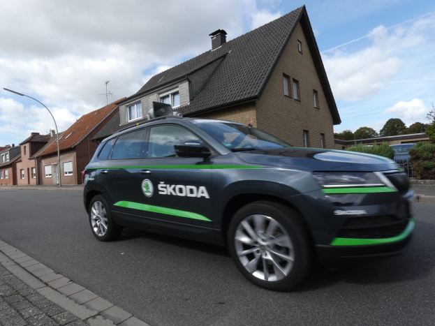 Schwarzer und grüner Skoda Kodiaq-Elektrowagen fährt auf einer Straße mit Gebäuden, Laternen, Bäumen und einem bewölkten Himmel.