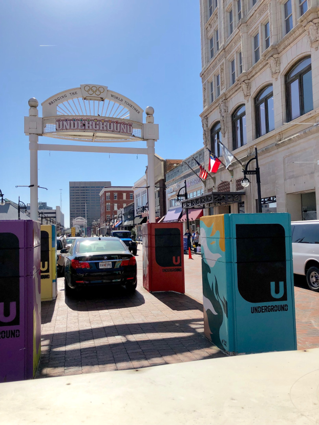 Stadtstraße mit parkenden Autos, ein dekorativer Bogen, Gebäude, Laternen, Fahnenmasten, ein Müllbehälter und Himmel im Hintergrund.