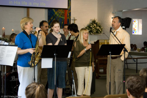 Eine Gruppe von Menschen in einem Raum mit einer Wandbanner, Kerzen, Mikrofonen, einem Fotorahmen und Blumen.