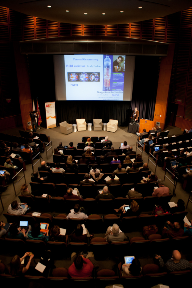 Ein mit sitzenden Menschen gefüllter Saal, der von vielen Lichtern beleuchtet wird, mit Fahnen auf der linken Seite, einem Podium auf der rechten Seite und einem Werbeplakat auf der linken Seite.