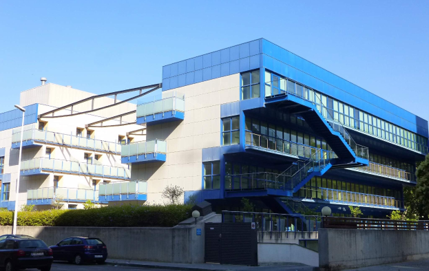 Großes beige Gebäude mit Reihen von parkenden Autos davor, umgeben von Straßenmasten, Laternen, Gittern, Grünflächen und einem klaren blauen Himmel, als das neue Hauptquartier der University of California, Berkeley bezeichnet.