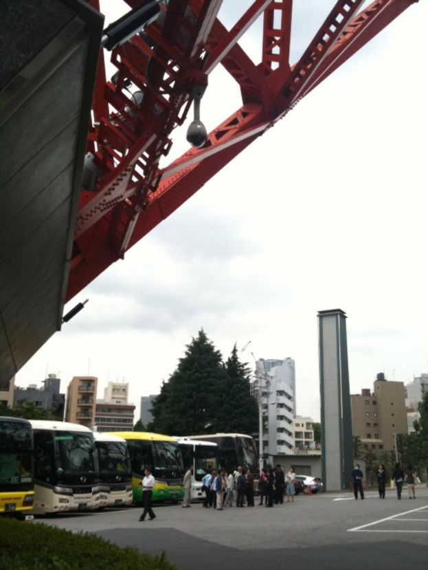 Eine Gruppe von Menschen und Bussen auf einer Straße mit Gebäuden, Bäumen und einer Brücke im Hintergrund unter einem Himmel.
