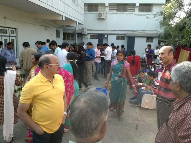 Menschen stehen, gehen und sitzen vor Gebäuden, einige halten Teller mit Essen in den Händen, mit einem Tisch voller Lebensmittel und Kleidung, die an einem Draht zur rechten Seite hängt, in der Nähe eines Baumes.