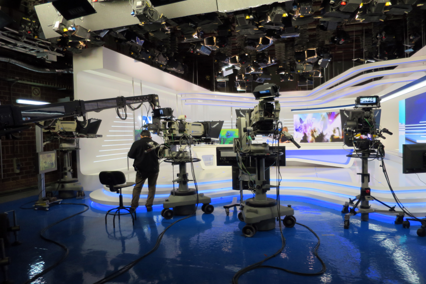 Ein Mann steht vor einem Fernseher und hält ein Mikrofon, umgeben von Kameras, einem Stuhl, einem Bildschirm, einer Wand und einer Decke mit Lampen, was auf eine Live-Übertragung hinweist.