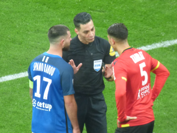 Schiedsrichter spricht mit zwei Männern auf einem Fußballfeld, einer trägt eine schwarze Hose und ein Hemd, der andere ein blau-rotes T-Shirt mit Text und Brille.