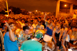 Menschen tanzen im Vordergrund bei einem Konzert mit Lichtern, Pfählen, Bäumen und anderen Objekten im Hintergrund.