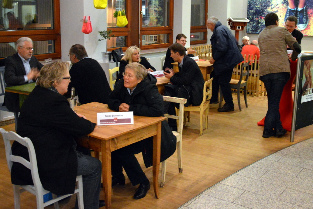 Eine Gruppe von Menschen, die an Tischen sitzen, einige stehen und sprechen, mit einer Tasche mit Pflanzen im Hintergrund.