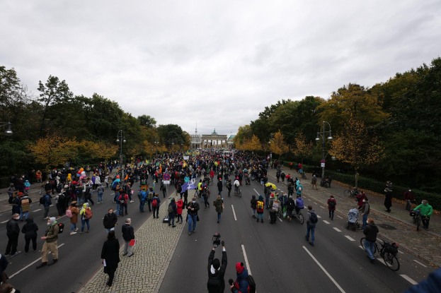 Eine große Gruppe von Menschen marschiert auf einer von Bäumen gesäumten Straße mit Laternenpfählen, hält Kameras, während sie an einer Protestdemo in Berlin teilnimmt, mit einem Gebäude und einem klaren Himmel im Hintergrund.