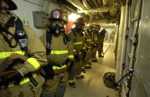 Eine Gruppe von Feuerwehrleuten in Helmen und Gasmasken geht einen Flur entlang, rechts eine Tür, Taschen auf dem Boden und Rohre und Lampen im Hintergrund.