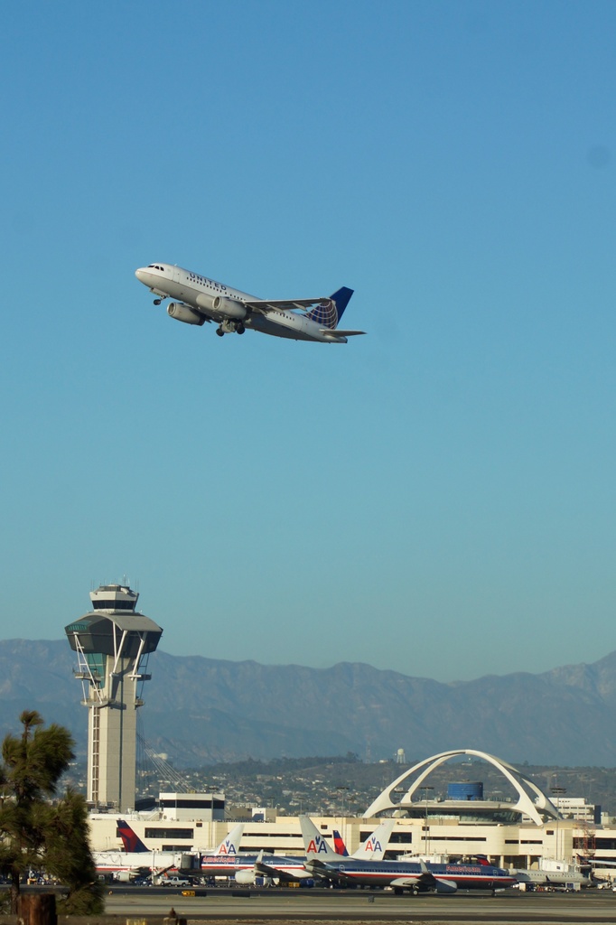 Ein Flughafenblick mit einer weißen Kontrollturm, geparkten Flugzeugen, fernen Gebäuden und einem Flugzeug am Himmel.