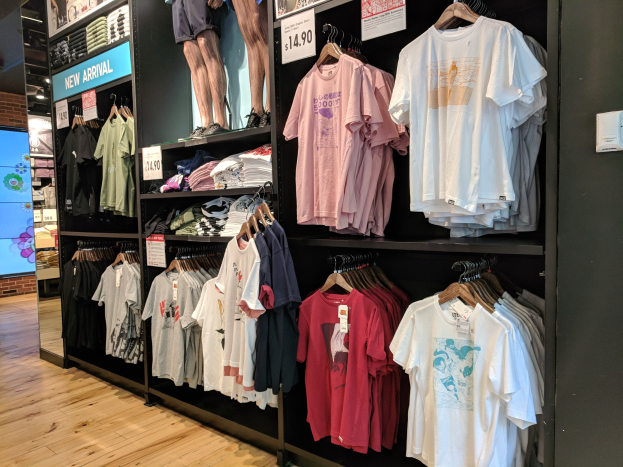 Eine Disney-Store-Ausstellung mit zahlreichen T-Shirts an Kleiderbügeln und Tafeln mit Texten und Bildern von Menschen, einem Bildschirm links und dem Boden unten.