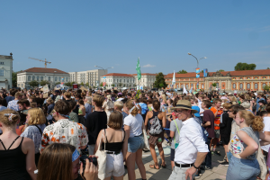 Eine große Menschenmenge steht vor einem Gebäude mit Fenstern, Laternenmasten, Flaggen und Bäumen, wobei einige Kameras halten, was auf eine Protest- oder Demonstrationsveranstaltung unter einem bewölkten Himmel hinweist.