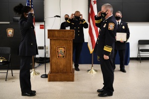 Gruppe uniformierter Personen in Masken vor einem Podium mit Mikrofon, umgeben von Flaggen und Möbeln, während einer Vereidigung, bei der eine Person ein Zertifikat hält.