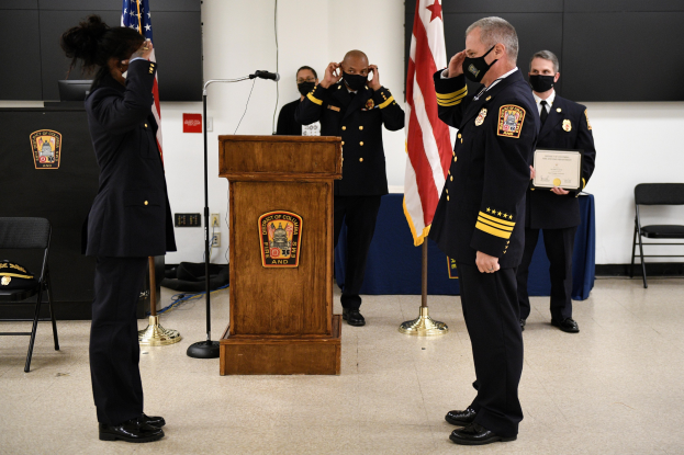 Gruppe uniformierter Personen in Masken vor einem Podium mit Mikrofon, umgeben von Flaggen und Möbeln, während einer Vereidigung, bei der eine Person ein Zertifikat hält.