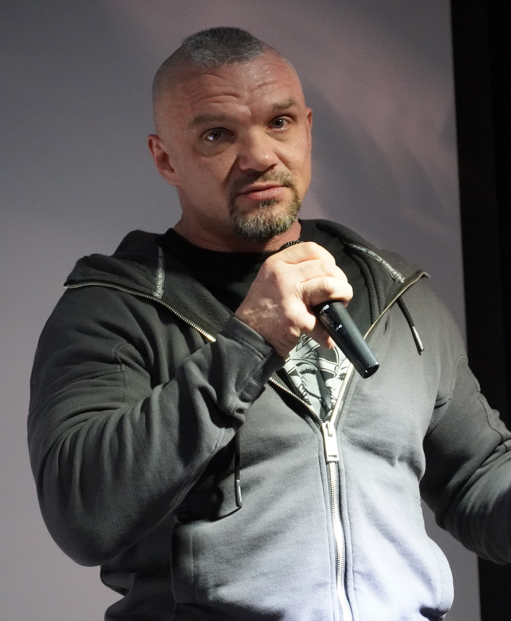 Ein Mann in einer schwarzen Kapuzenjacke und einem schwarzen Anzug mit einem wei├čen Hemd, der ein Mikrofon h├Ąlt, steht selbstbewusst vor einer Wand und scheint ein Kom├Âdiant zu sein.