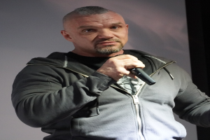 Ein Mann in einer schwarzen Kapuzenjacke und einem schwarzen Anzug mit einem wei├čen Hemd, der ein Mikrofon h├Ąlt, steht selbstbewusst vor einer Wand und scheint ein Kom├Âdiant zu sein.