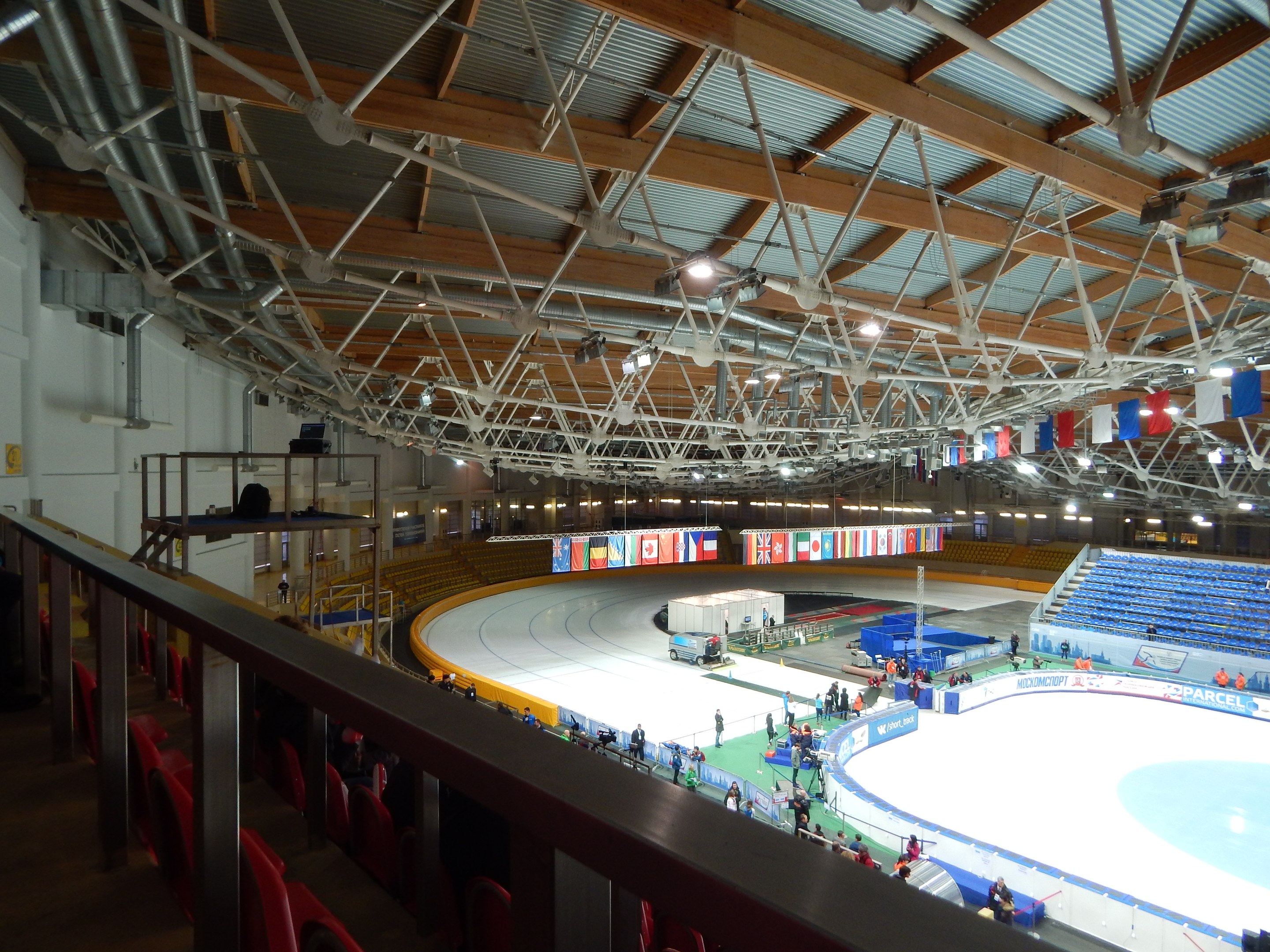Große Indoor-Eisfläche mit zahlreichen Menschen, die Schlittschuh laufen, umgeben von Sitzplätzen, Geländern, Brettern, Beleuchtungskörpern und strukturellen Elementen, mit einer Wand im Hintergrund.