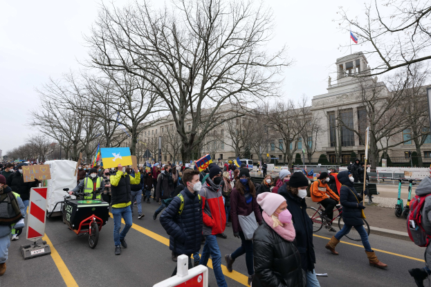 Eine große Gruppe von Menschen bei einer Demonstration auf einer Straße, einige halten Schilder und andere fahren Fahrräder, mit Bäumen und einem klaren blauen Himmel im Hintergrund.