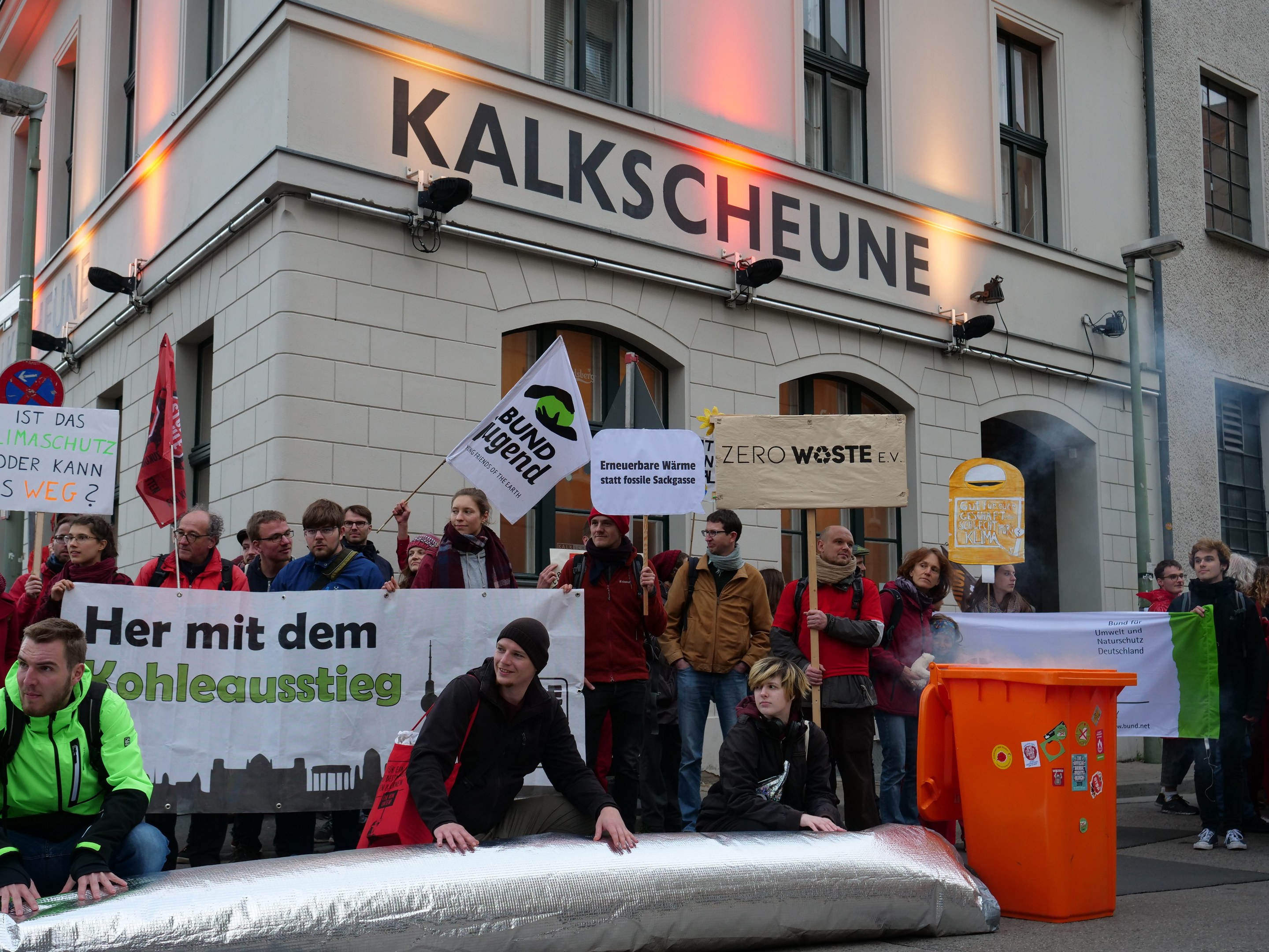 Eine Gruppe von Menschen mit Schildern und Transparenten vor einem Gebäude während einer Demonstration in Deutschland, mit zwei Personen im Vordergrund und einem Müllcontainer auf der rechten Seite.