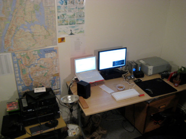 Ein Raum mit einem Tisch, auf dem ein Laptop, Lautsprecher, Monitor, Tastatur, Ball, Maus, Scanner und Papiere liegen, und einem weiteren Tisch mit Elektronikgeräten im Hintergrund, mit Charts und Postern an der Wand dahinter.