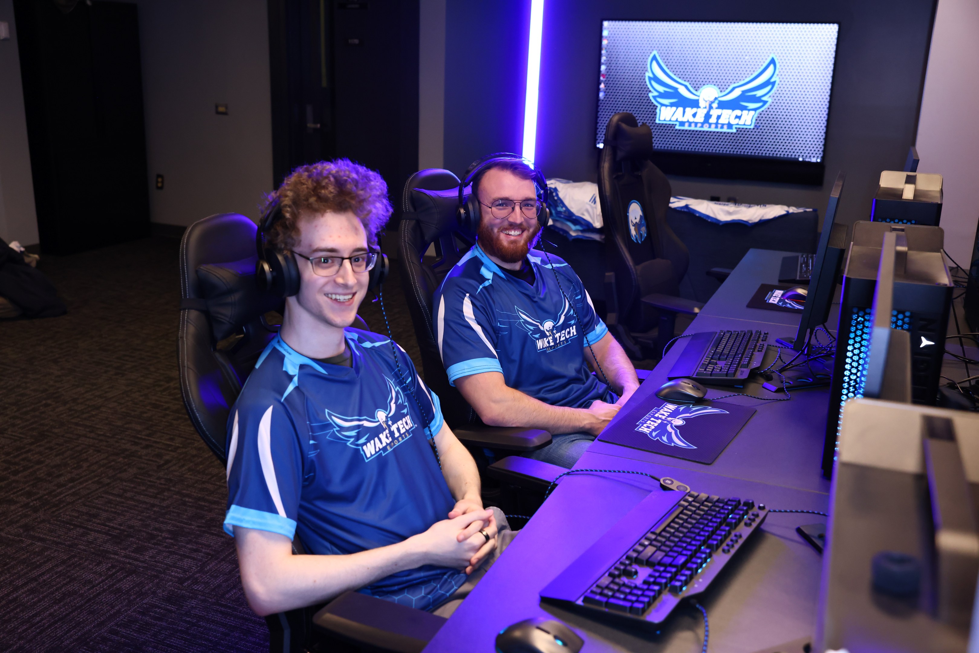 Zwei Männer mit Headsets sitzen an einem Schreibtisch mit Monitoren, Tastaturen und anderem Equipment und lächeln, während sie ein Spiel der Wake Tech League of Legends spielen.