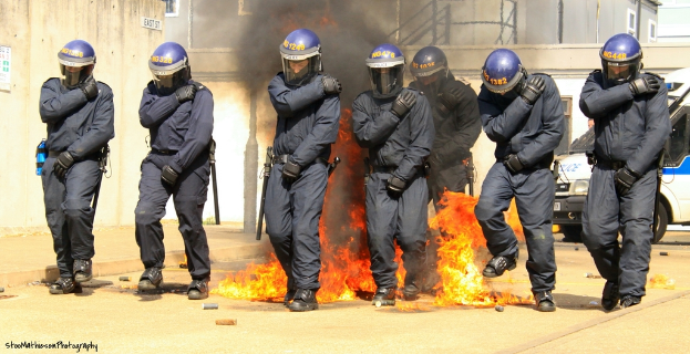 Menschen in Helmen stehen vor einem Feuer mit verstreuten Gegenständen, Gebäuden und einem Fahrzeug im Hintergrund, einem Plakat und einer Tafel an der linken Wand und Text am unteren Rand des Bildes.