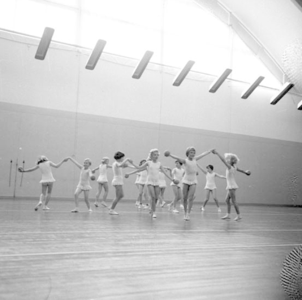 Schwarzes und weißes Bild von jungen Mädchen, die in einem Studio tanzen und sich an den Händen halten, mit einem Ball auf der rechten Seite und Lampen an der Decke; ein Wandobjekt ist im Hintergrund sichtbar.