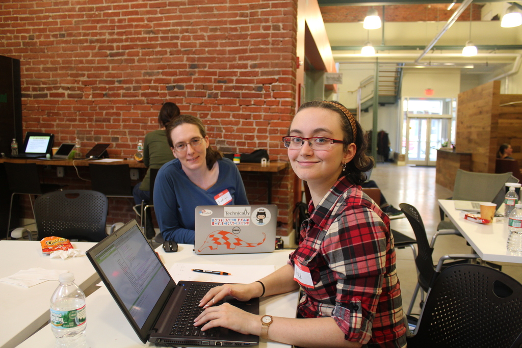 Zwei Frauen an einem Tisch mit Laptops, einer Flasche, einem Stift und Papier in einem Büro mit einer roten Backsteinwand, Deckenlampen, einer Treppe und einer Eingangstür im Hintergrund.