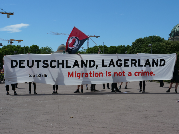 Eine Gruppe von Menschen mit einem Transparent mit der Aufschrift "Deutschland, Lagerland Migration ist kein Verbrechen" und einer Flagge, im Hintergrund Straßenlaternen, Bäume, Kräne, ein Gebäude und ein bewölkter Himmel.