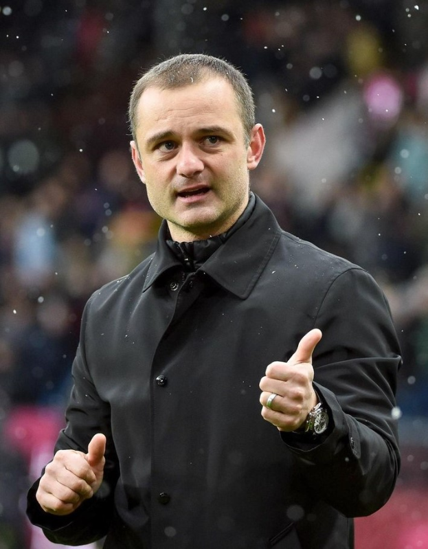 Ein Mann in einer schwarzen Jacke zeigt den Daumen hoch, trägt eine Uhr und hat einen unscharfen Hintergrund.