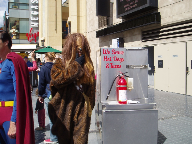 Eine Menschenmenge, einige in Kost√ľmen, versammelt sich vor Geb√§uden mit sichtbaren Schildern, einer Box mit Schrift und einem Feuerl√∂scher.