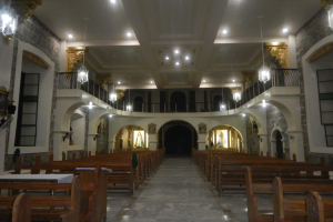Das Innere einer Kirche mit hölzernen Kirchenbänken, Tischen in der Mitte, Geländern, Lampen und Skulpturen, mit detailreichen Wandmalereien und einer warm beleuchteten Decke.