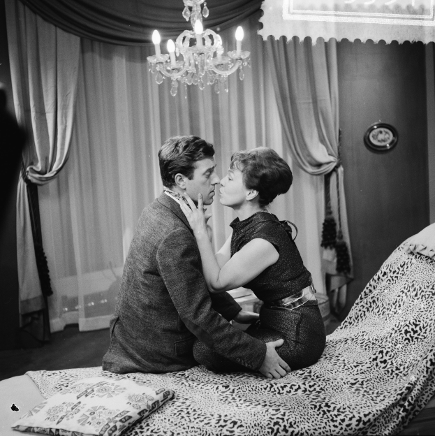 Ein Mann und eine Frau auf einem Bett sitzend, die sich umarmen und küssen, mit Kissen und einer Decke auf dem Bett, einem Kronleuchter an der Decke und Vorhängen im Hintergrund.