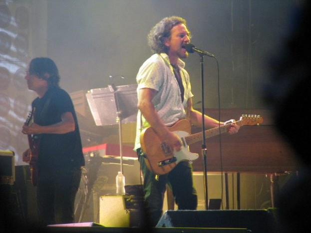 Ein Live-Musikauftritt auf der Buhne mit einem Mann in weissem Hemd, der in ein Mikrofon singt und Gitarre spielt, begleitet von einem anderen Mann in schwarzem Hemd, der ebenfalls Gitarre spielt, mit einem Piano, Kabeln, Lautsprechern und Buhnenlichtern im Hintergrund.