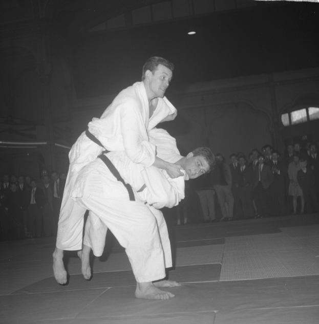 Zwei Männer in Judo-Uniformen rangeln in der Mitte eines Schwarz-Weiß-Bildes, mit einer Gruppe von Menschen und Lichtern im Hintergrund.
