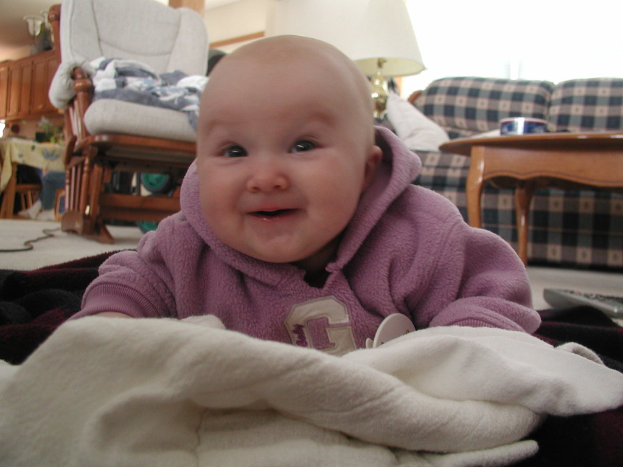 Ein Junge lächelt drinnen mit einem Stuhl, Sofa, Tisch, Lampe, Kleiderschrank und einem weiteren Tisch im Hintergrund, während ein Kabel und Decken auf dem Boden um ihn herum liegen.