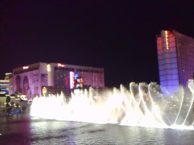 Ein Wasserbrunnen in der Mitte mit Wasser am Boden, umgeben von Gebäuden, Plakaten, Bildschirmen, Lichtern, Pflanzen und Himmel im Hintergrund.
