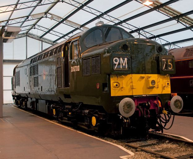 Ein Zug fährt auf den Schienen neben einem Bahnsteig mit einem Mast auf dem Dach, der Metallstangen mit Lampen trägt.