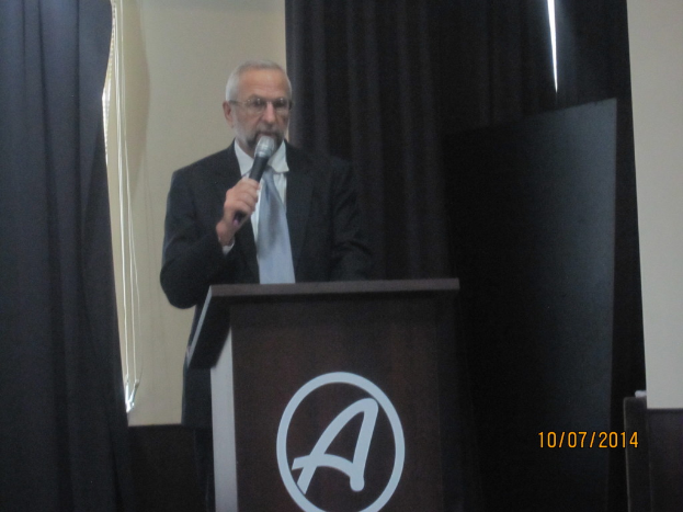 Eine Person in einem schwarzen Mantel und grauem Schlips steht an einem Schreibtisch und hält ein Mikrofon, während sie eine Rede hält, mit schwarzen Vorhängen im Hintergrund.