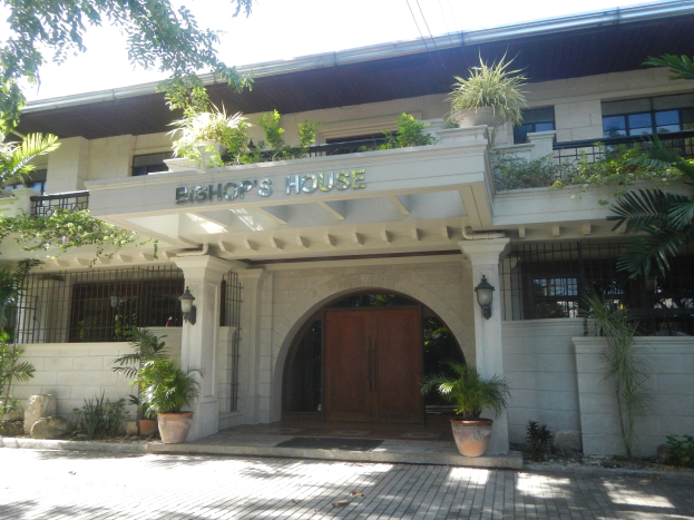 Das Bischofshaus in Manila, Philippinen, mit Fenstern, einer Tür, einem Namensschild, Topfpflanzen, umliegenden Bäumen, verstreuten Steinen und einem bewölkten Himmel.