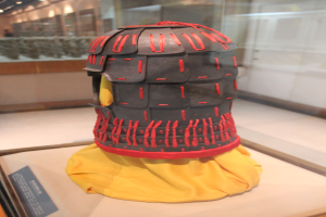 Ein Helm in einer Glasvitrine mit einem gelben Tuch darüber, begleitet von einer Texttafel auf der linken Seite und Deckenlampen im Hintergrund.